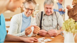 An elderly couple with a caregiver engaged in a jigsaw puzzle, fostering connection and cognitive engagement amidst dementia and Alzheimer’s challenges.