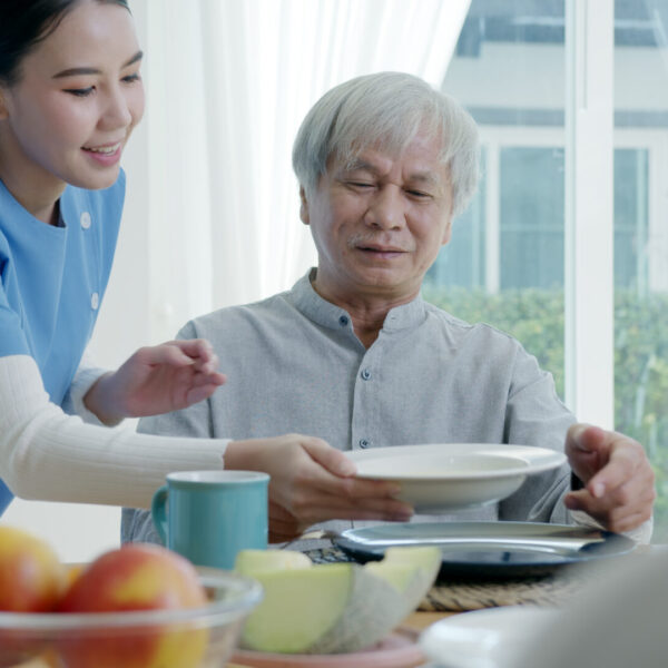 A caregiver is serving food to an elderly person with Alzheimer’s disease, while there is another person facing the elderly individual
